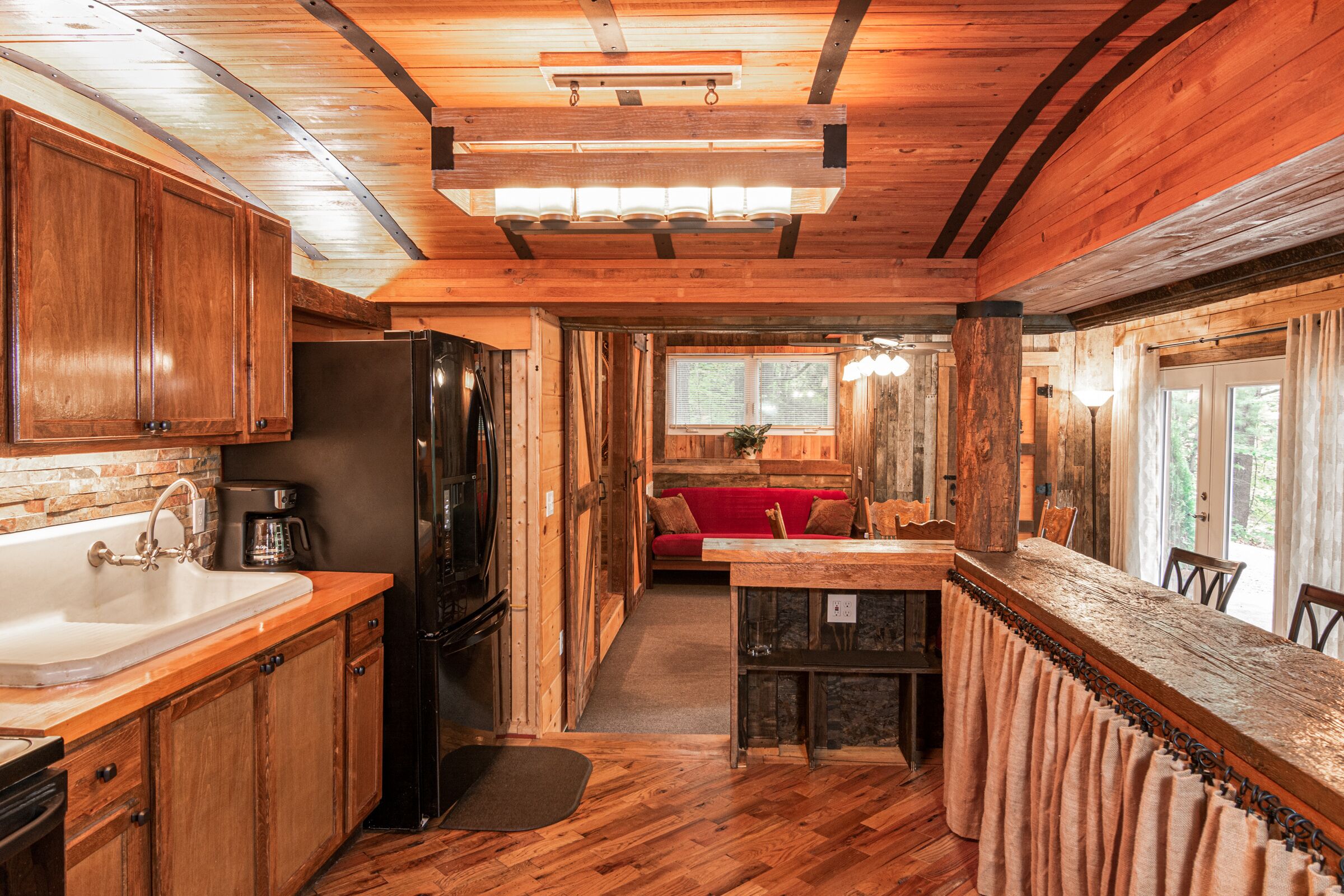 A rustic cabin interior with wooden floors, a kitchen with a sink and refrigerator, a dining area with a table and chairs, and a cozy seating area with a red sofa.