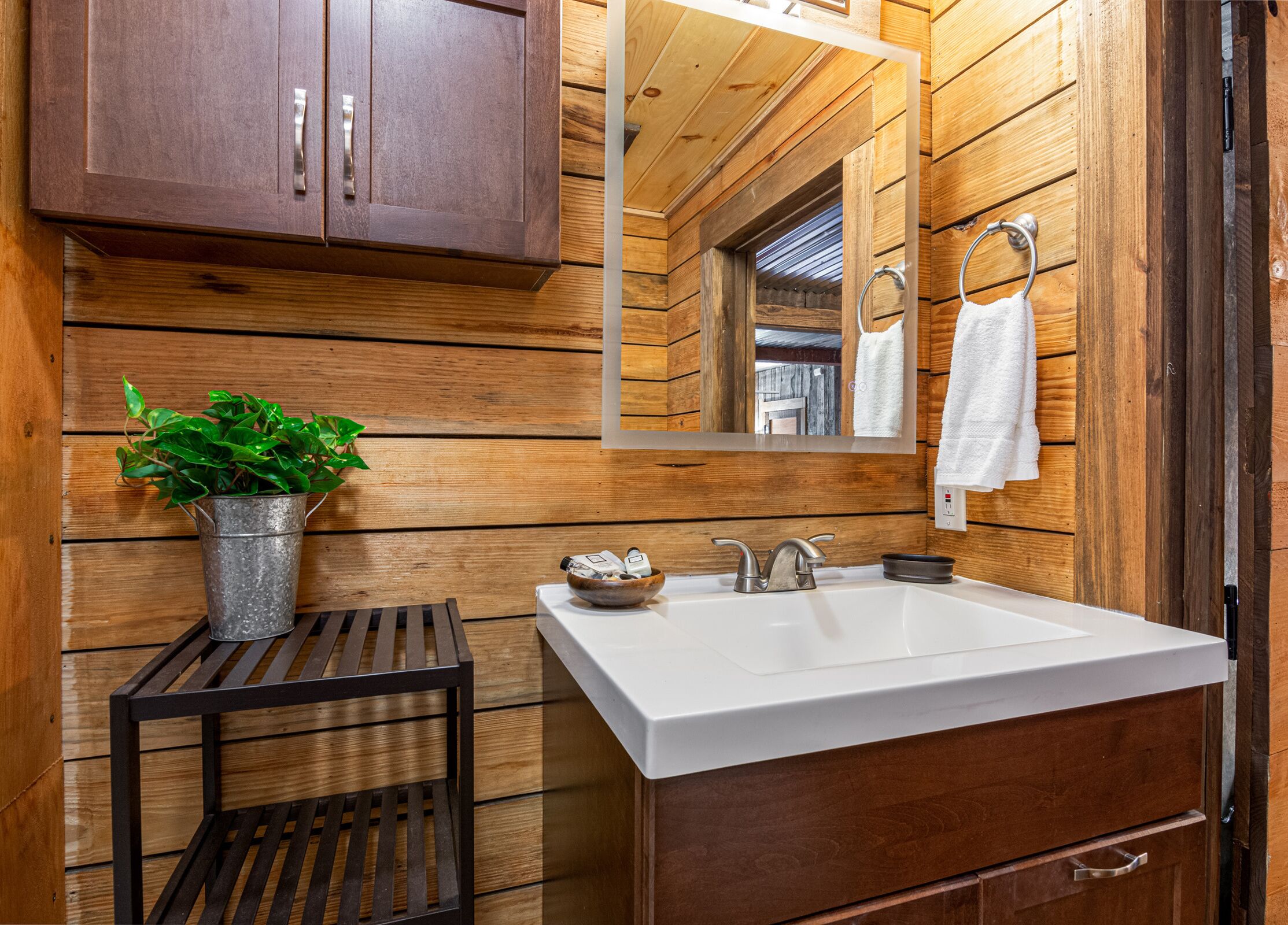 A wooden cabin bathroom with a white sink, wooden cabinets, a mirror, and a potted plant.