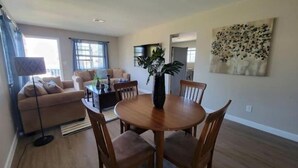 Un salon avec un canapé, une table basse et une table à manger avec des chaises. On y trouve une lampe, une plante et un tableau accroché au mur.