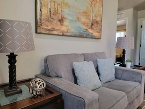 Un salon avec un canapé gris, des lampes à motifs, une sphère décorative et un tableau représentant une scène de forêt.