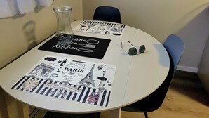 A round dining table with black placemats featuring Parisian illustrations, a clear glass vase, and a pair of sunglasses. A painting of a street scene hangs on the wall.