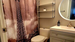 A bathroom with a shower curtain featuring the Eiffel Tower, a toilet, a sink with a mirror, and a shelf with a small plant.