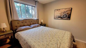 A bedroom with a large bed, a nightstand with a lamp, a window with curtains, and a wall-mounted bicycle artwork.