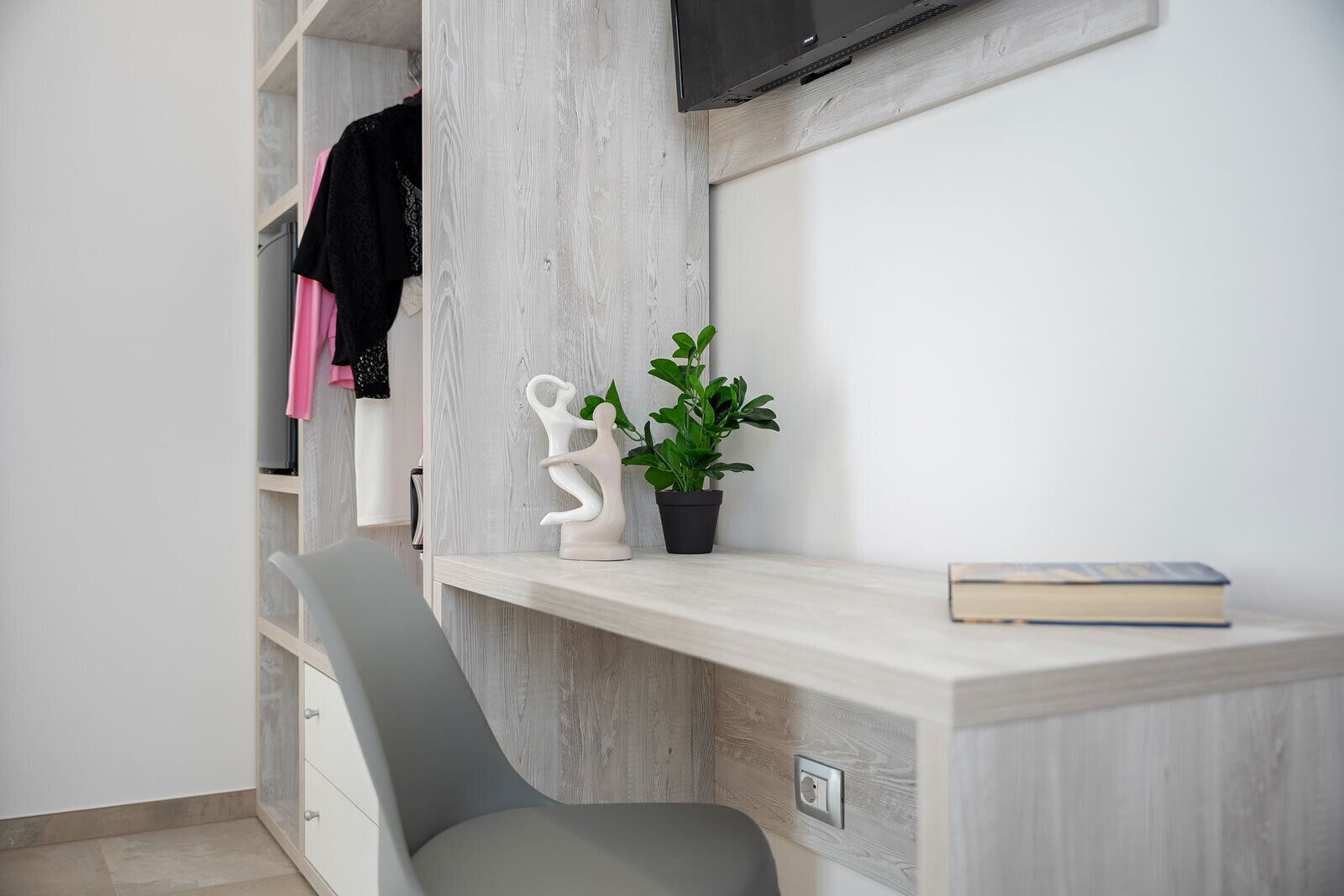 Una habitación moderna con escritorio empotrado, una silla, televisor montado en la pared y una planta en maceta.