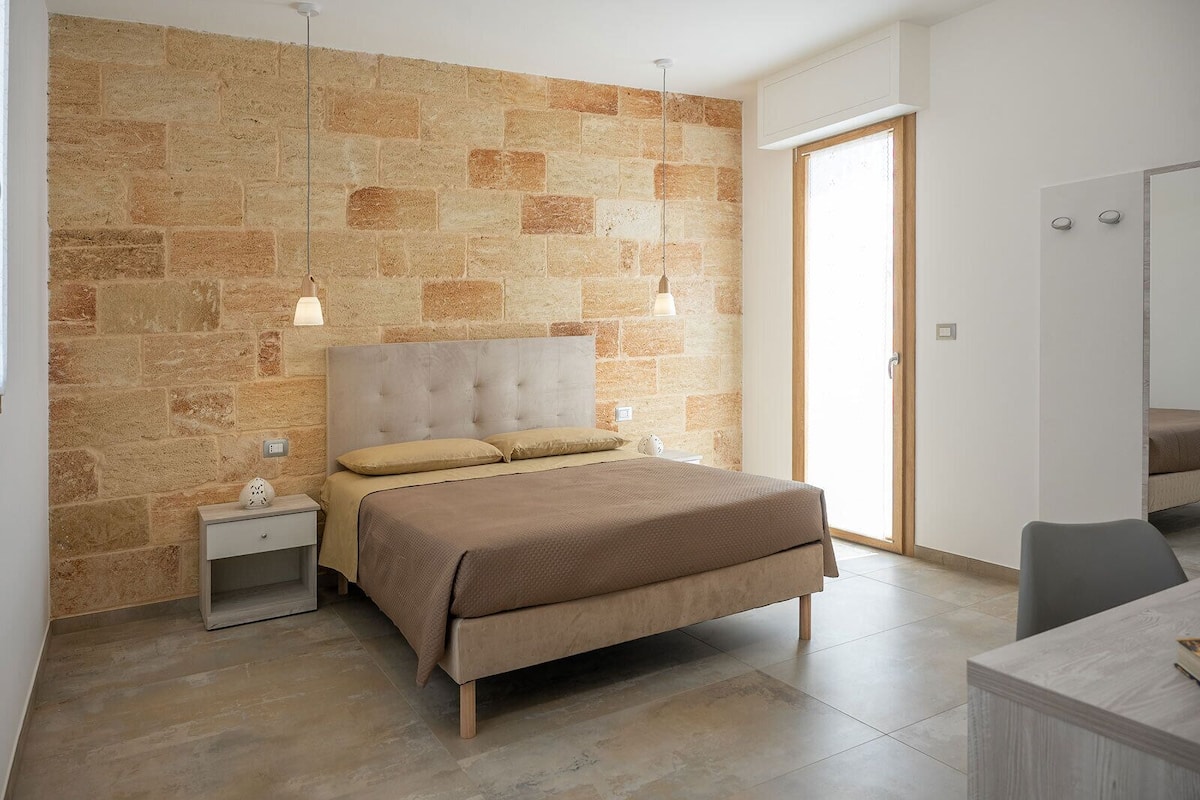A bedroom with a stone wall, a bed with beige bedding, a nightstand, a desk, and a chair.