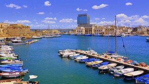 Uma marina com vários barcos atracados, um edifício imponente ao fundo e um céu azul claro.