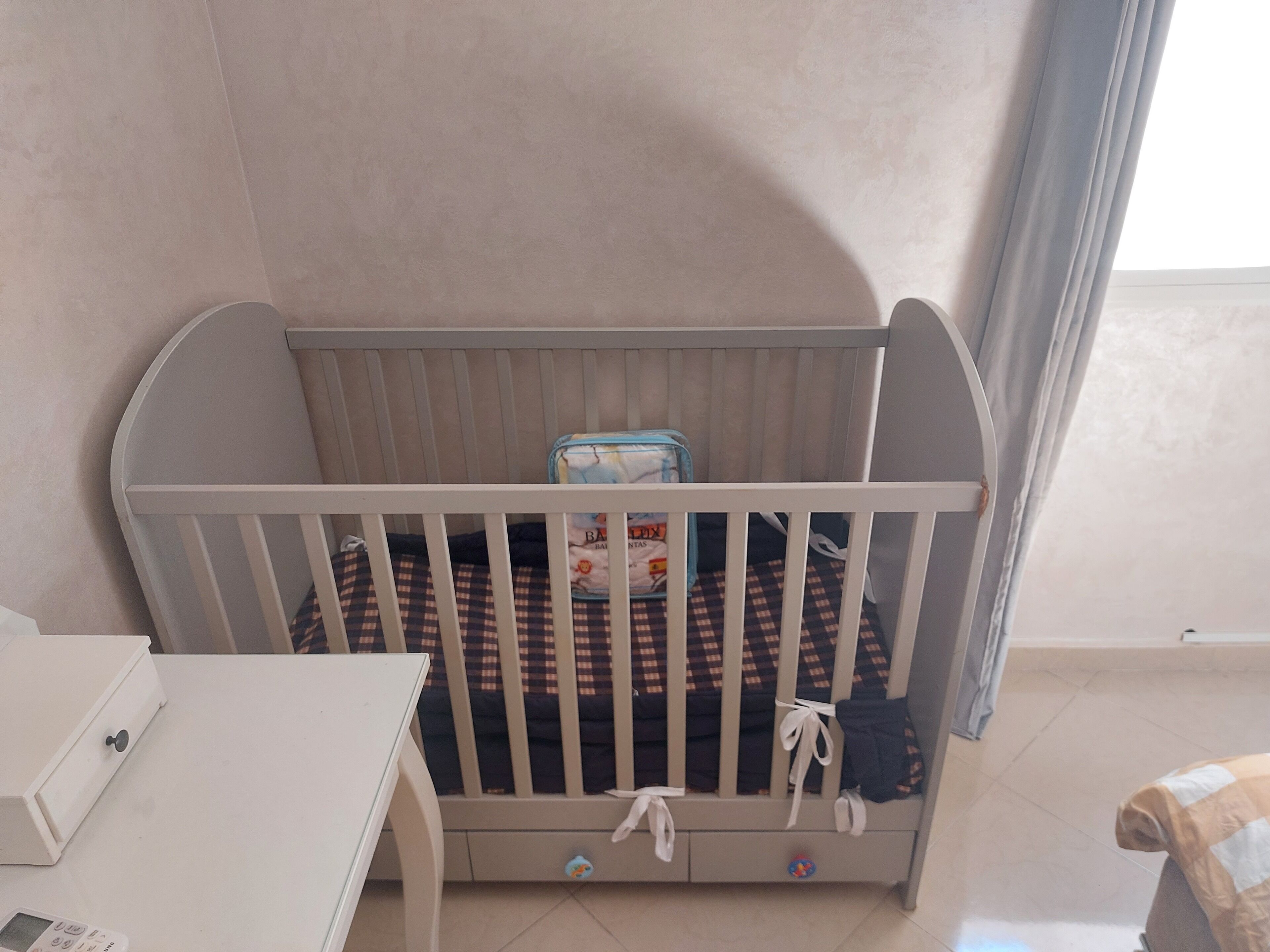 Un berceau blanc avec un matelas à carreaux, un petit bac de rangement et un jouet dans le coin.