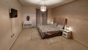 A bedroom with a bed, bedside table, lamp, dresser, mirror, and a television mounted on the wall. - Appartement Centre-ville (Kénitra)