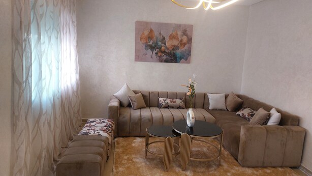 A living room with a sofa, ottoman, and a round table with chairs. There is a painting on the wall and a vase with flowers on the table. - Appartement Centre-ville (Kénitra)