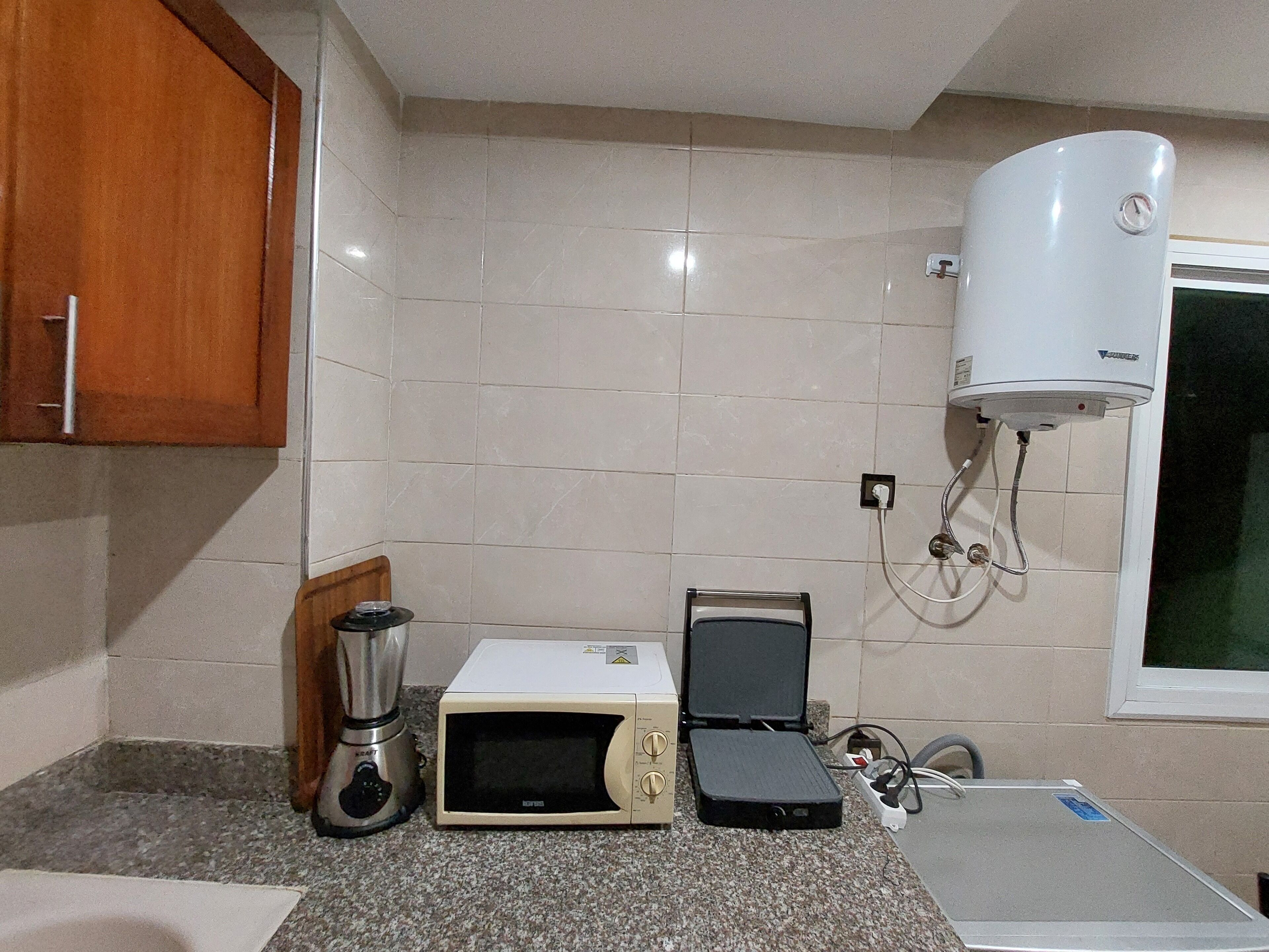 Una cocina con microondas, tostadora y licuadora sobre la encimera, un calentador de agua instalado en la pared y una ventana con vistas al exterior.