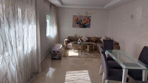 A living room with a sofa, ottoman, glass coffee table, and a dining table with chairs. - Appartement Centre-ville (Kénitra)