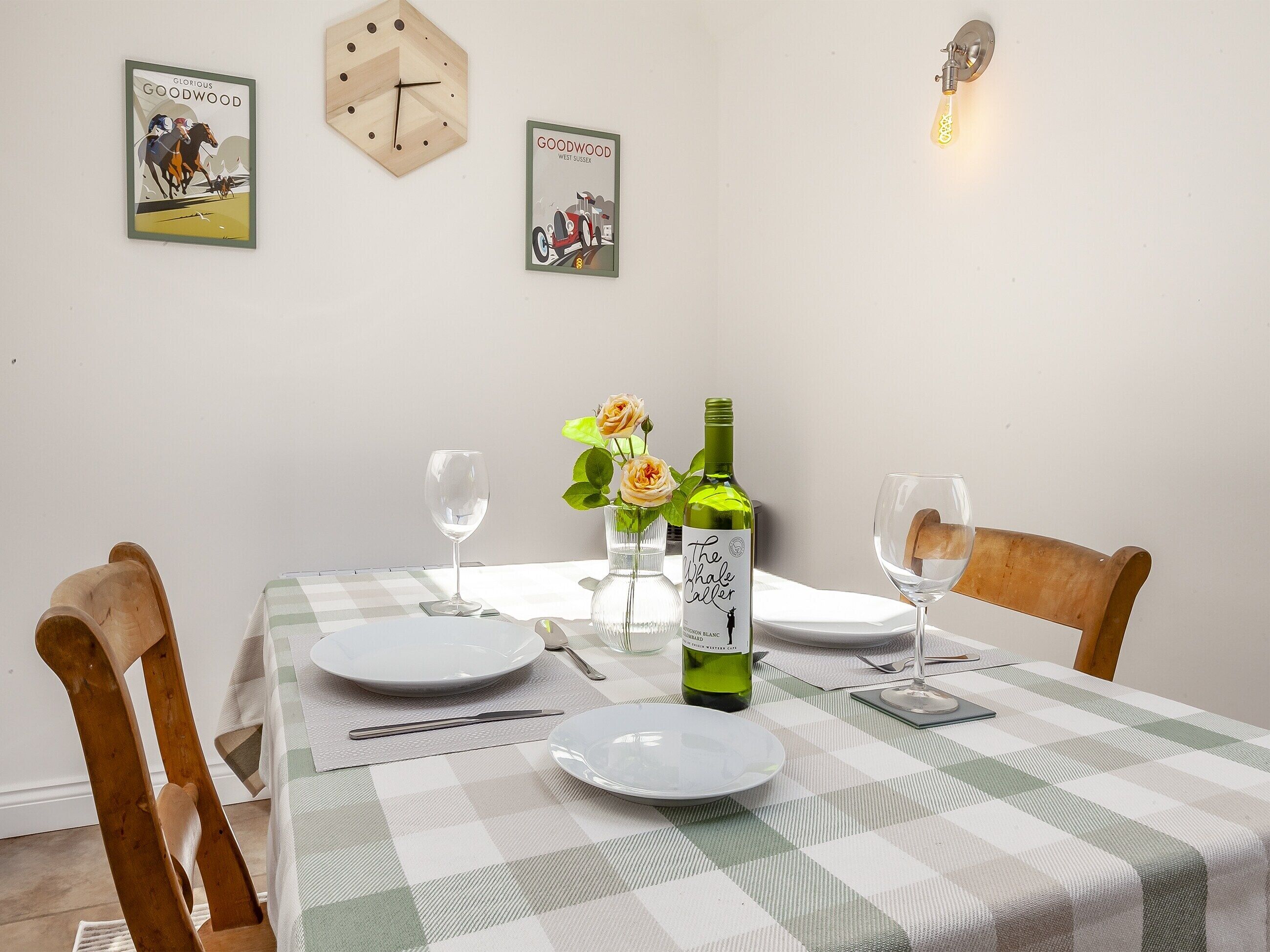 Une table à manger dressée avec des assiettes, une bouteille de vin et des verres, ainsi qu’une horloge et des affiches accrochées au mur.