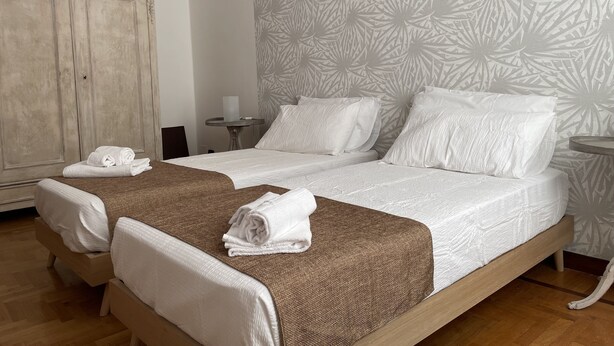 Una cama doble con sábanas blancas y una manta marrón, un armario de madera y un papel tapiz estampado.