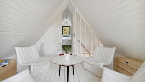 A white, sloped-roofed room with two white chairs and a small table, a potted plant, and a window with a view.