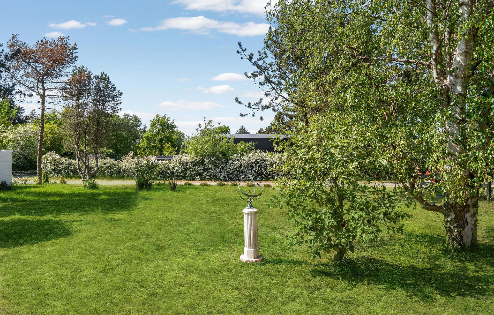 A well-maintained garden with a sundial, various trees, and a building in the background.