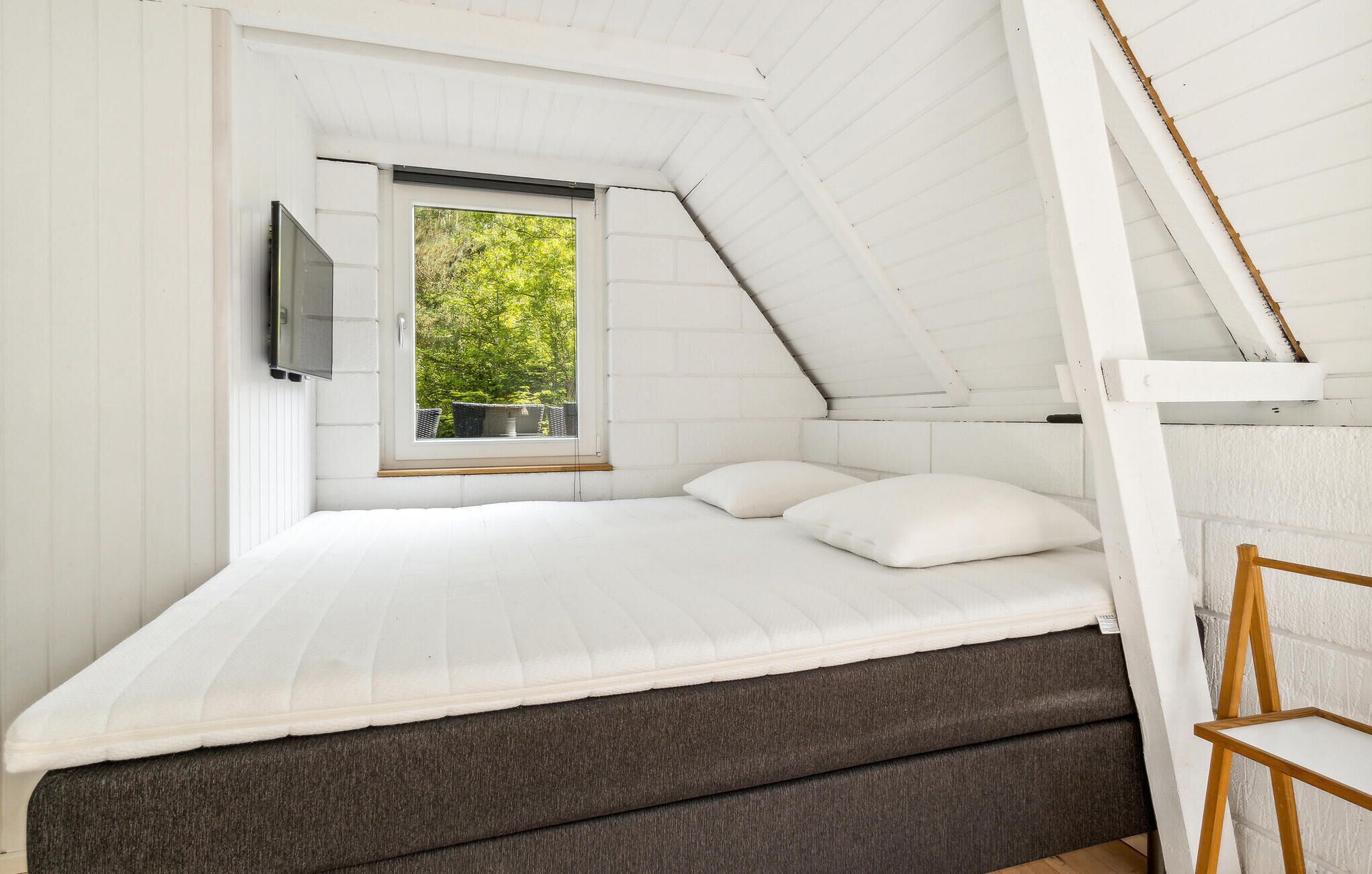 Une chambre avec un plafond en pente, une fenêtre donnant sur la verdure, un lit avec une literie blanche et une échelle en bois.