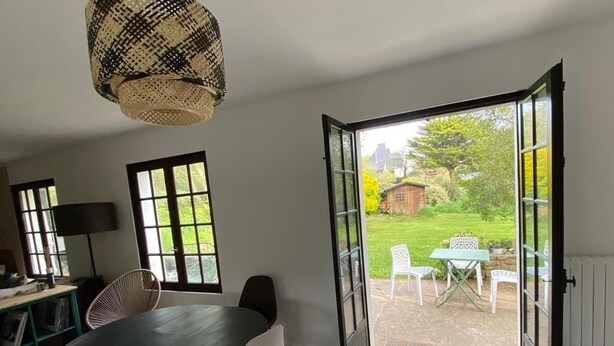 Una puerta abierta que da a un jardín con una mesa y sillas, una mesa negra en el interior y una lámpara de mimbre.