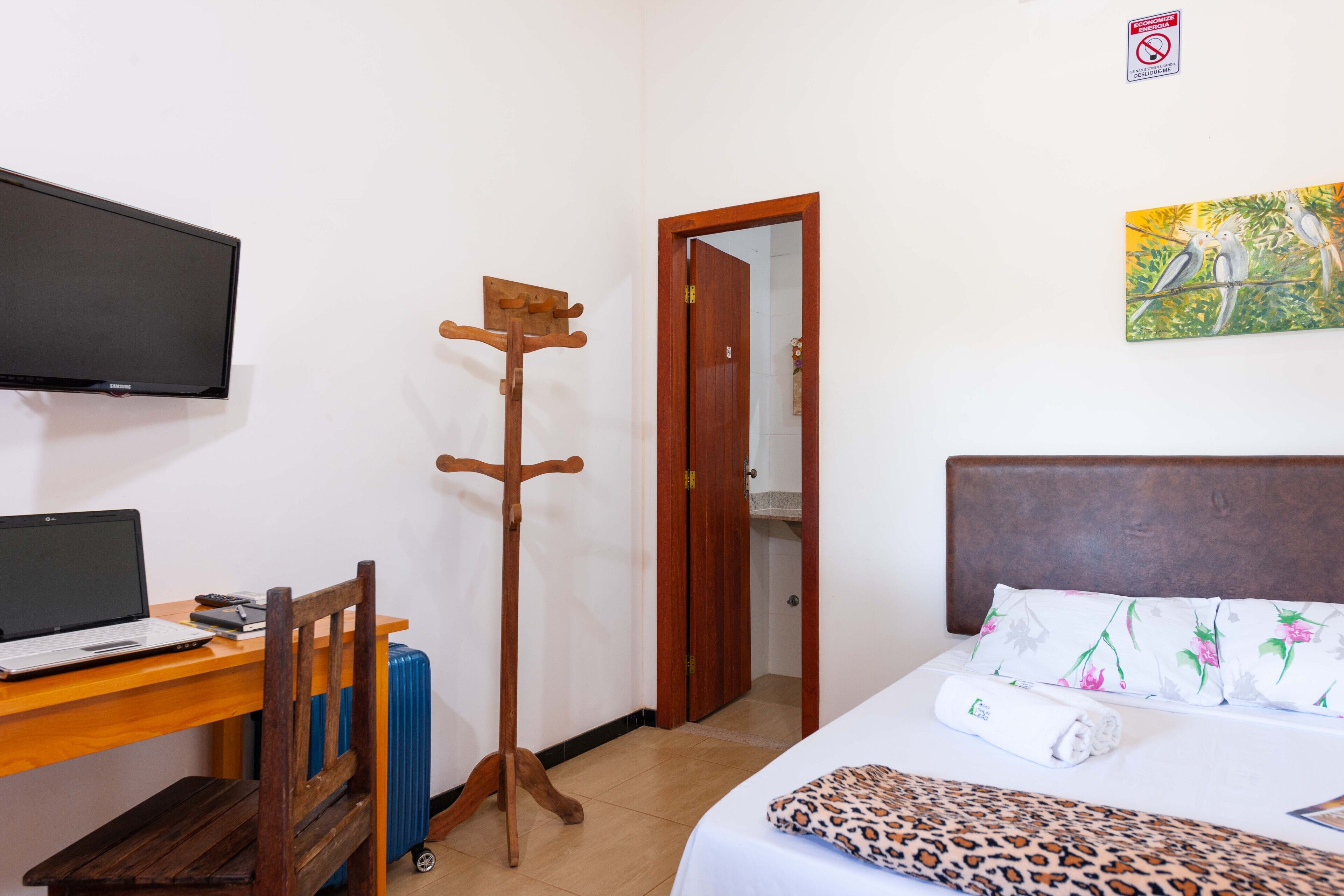 A hotel room with a bed, a desk with a laptop, a coat rack, a TV, and a painting on the wall.