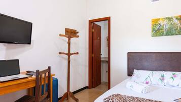 A hotel room with a bed, a desk with a laptop, a coat rack, a TV, and a painting on the wall.