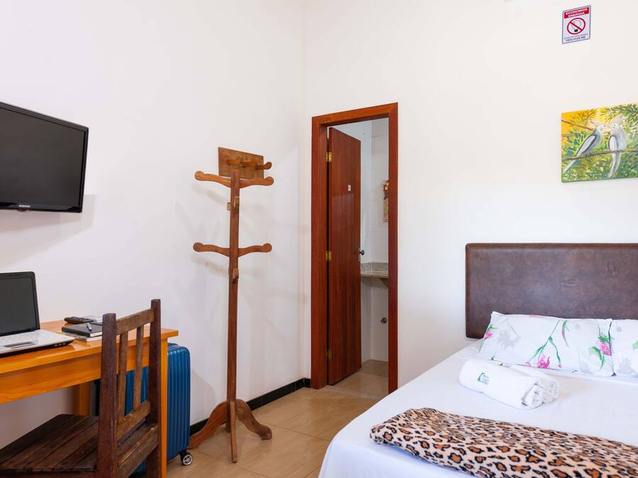 A hotel room with a bed, a desk with a laptop, a coat rack, a TV, and a painting on the wall.