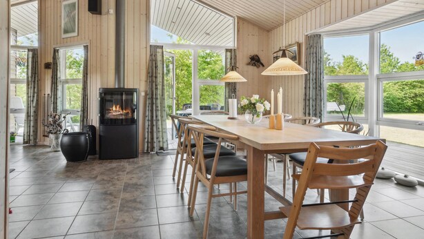 A spacious dining area with a long wooden table and chairs, a black wood-burning stove, and large windows offering views of greenery.