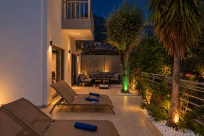 An outdoor patio area with lounge chairs, a dining table, and a decorative tree illuminated by green lights.