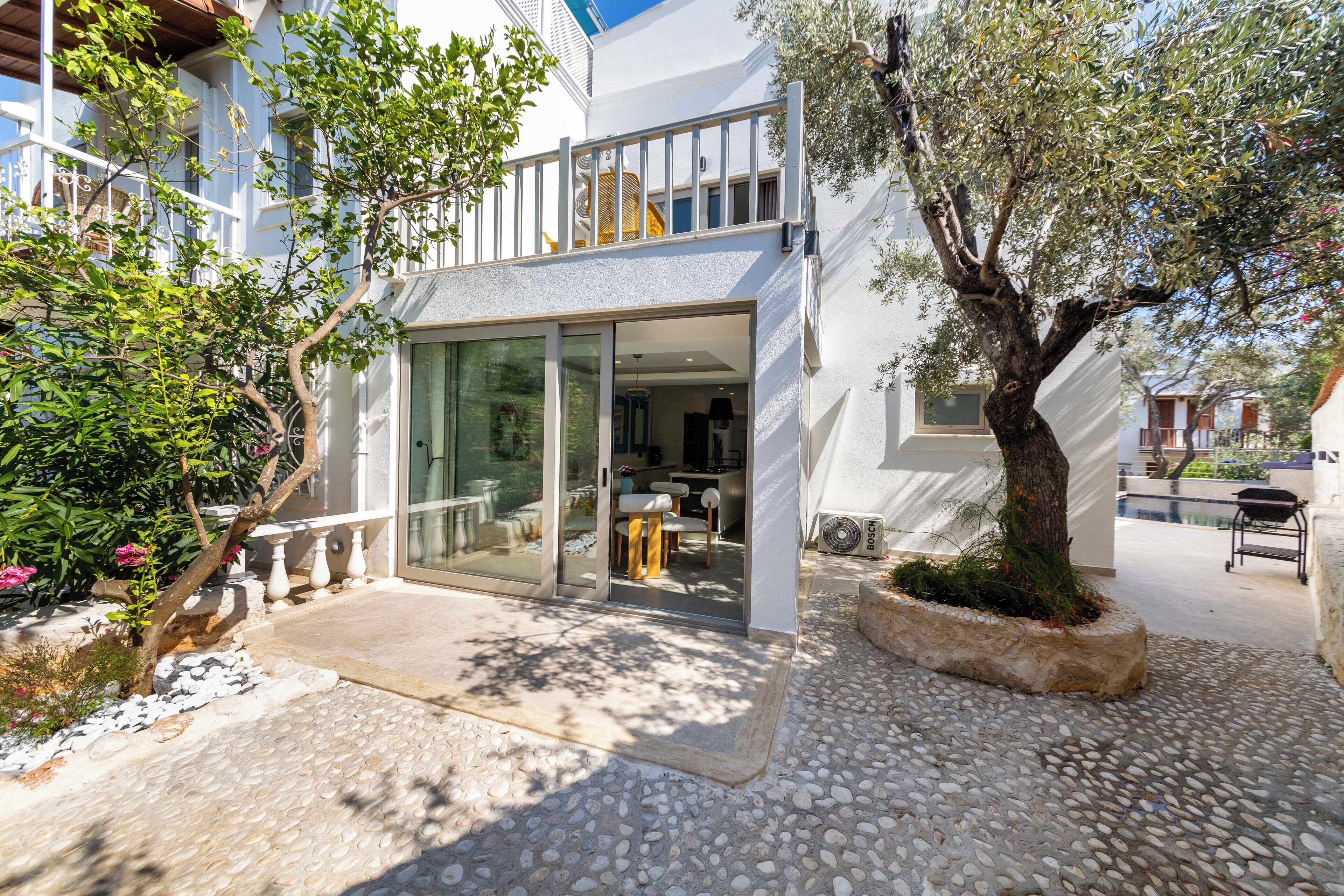 A modern house with a glass door entrance, a balcony, and a garden area with a tree and plants.