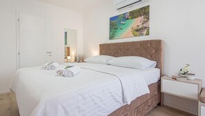 Ein ordentlich bezogenes Bett mit weißen Leinen und gerollten Handtüchern, ein gepolstertes Kopfteil, ein Nachttisch mit Lampe sowie ein Strandbild über dem Bett.