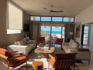 A spacious living room with a large window overlooking the beach, a ceiling fan, and a dining area with outdoor seating. - Beautiful Beach front Villa (La Ribera, Baja California Sur, Mexico)