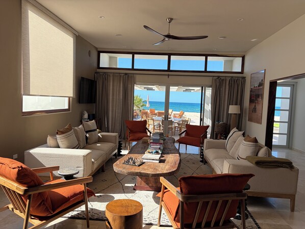Un salón amplio con un ventanal grande con vista a la playa, un ventilador de techo y una zona de comedor con mobiliario de exterior.