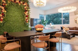 A modern living room with a large dining table, comfortable seating, a decorative wall, and a chandelier.