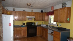 A kitchen with wooden cabinets, a stove, a refrigerator, and a window with a red curtain. - Rocking J Ranch House (Cove)