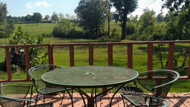 A wooden deck with a green metal table and chairs, overlooking a grassy landscape with trees. - Rocking J Ranch House (Cove)
