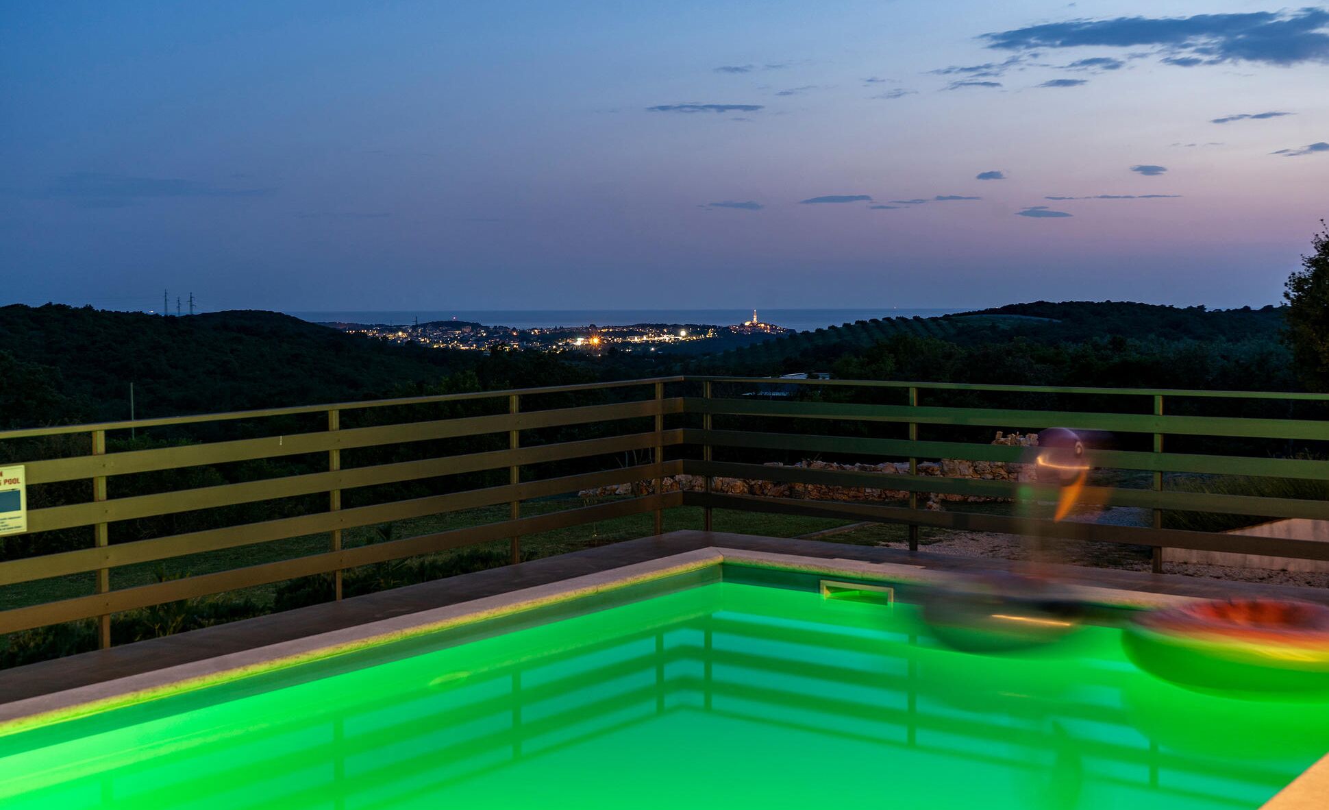 A swimming pool with a wooden fence and a view of a distant cityscape at dusk.
