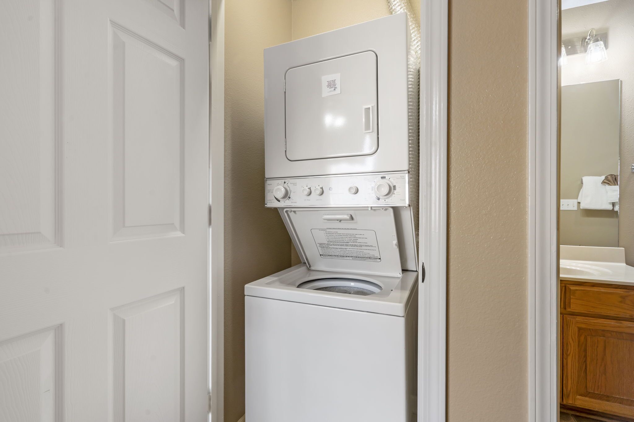 Een compacte wasmachine en droger staan in een kleine wasruimte, met een deur en een spiegel die een deel van de ruimte weerspiegelt.