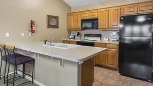 Una cocina con armarios de madera, un refrigerador negro, un microondas, un lavamanos y un taburete de bar.