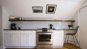Una cocina con armarios blancos, horno de acero inoxidable y encimera de madera. Hay dos cuadros en la pared y una silla en la esquina.