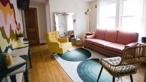 Una sala de estar con un sillón amarillo, un sofá rosa, una silla de mimbre, un espejo y una lámpara de pie.