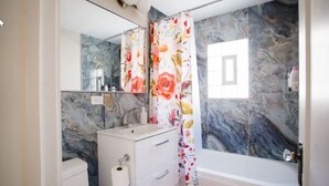 Un baño con pared de mármol, un lavamanos blanco, inodoro y cortina de ducha con motivos florales.
