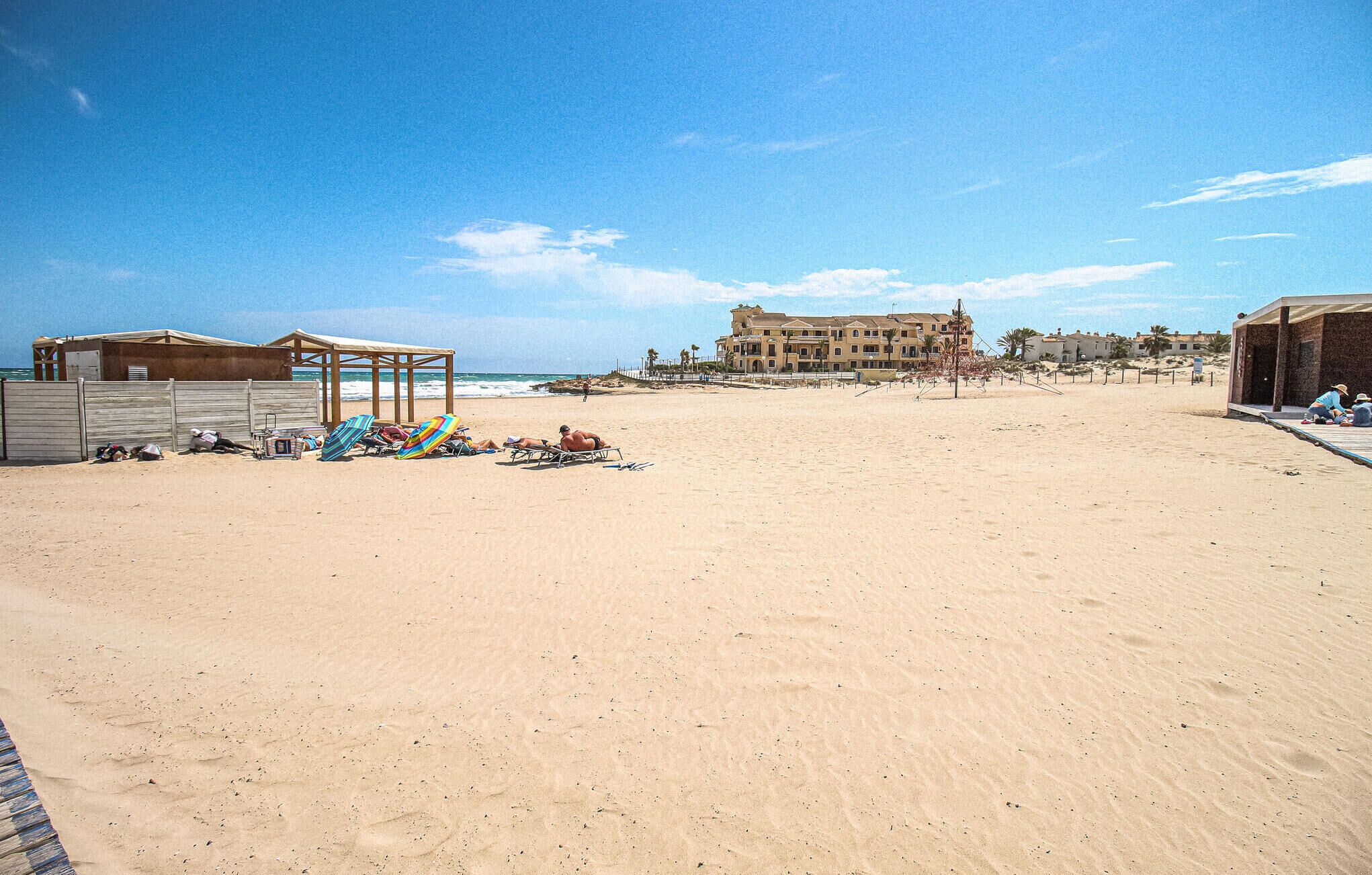 A beach with sun loungers and umbrellas, a building with multiple balconies, and a clear sky.