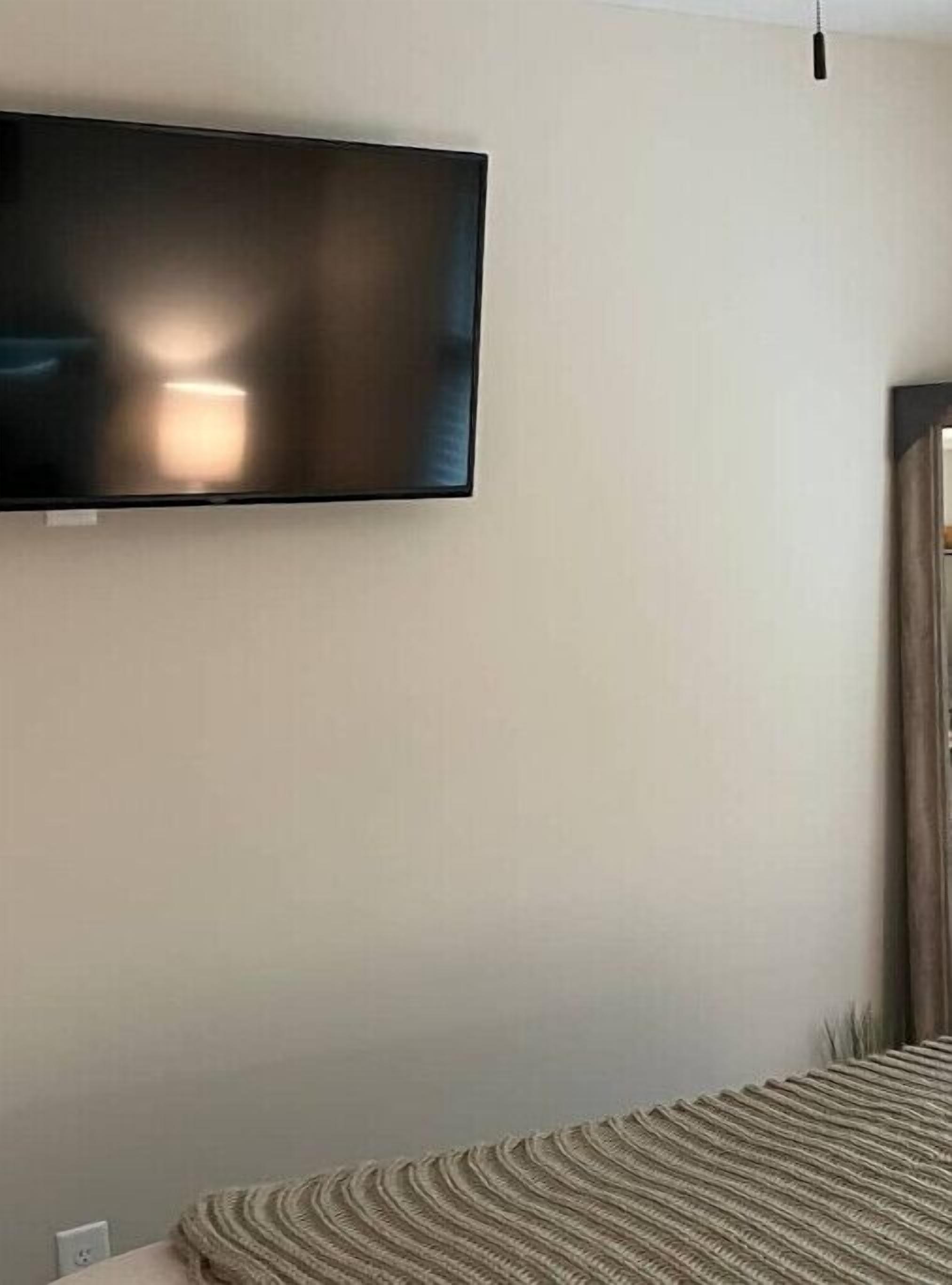 Ein Zimmer mit einem flachbildfernseher an der Wand, einem Bett mit gestreifter Bettdecke und einem Fenster mit Vorhängen.