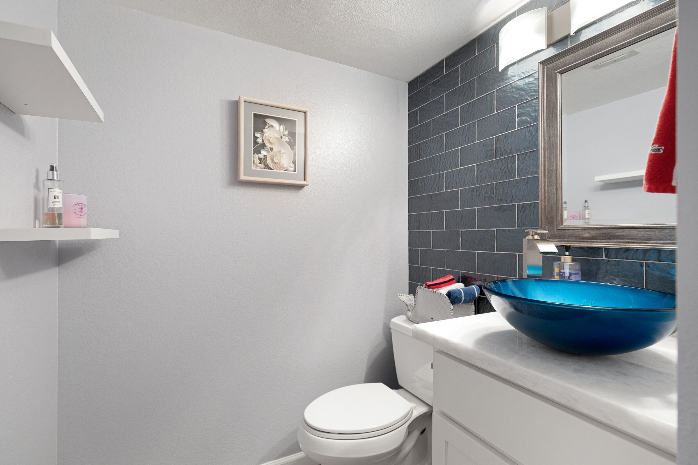 A bathroom with a white toilet, a blue basin sink, a framed picture on the wall, and a red towel hanging on the mirror.