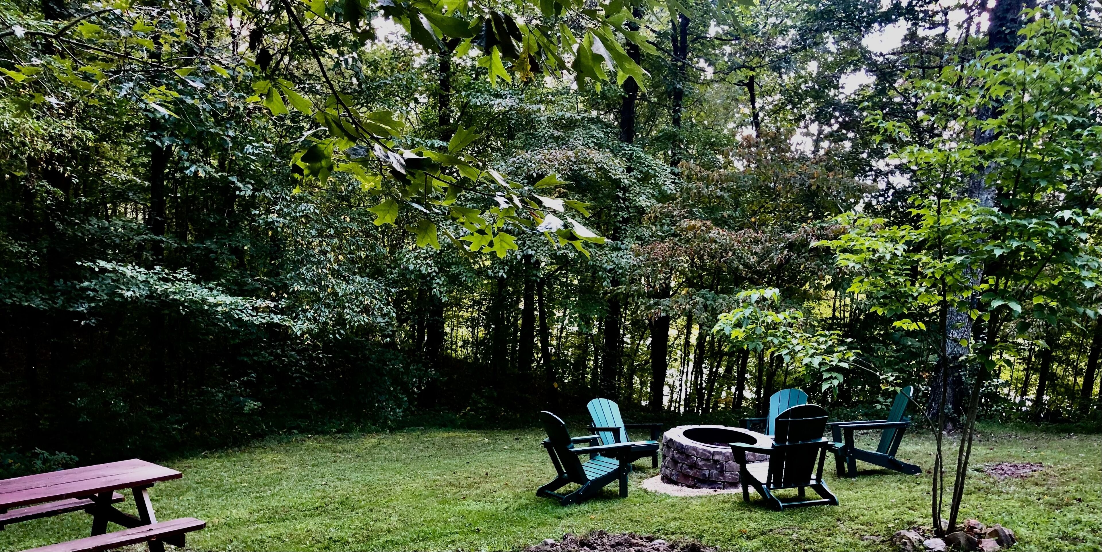 A backyard with a stone fire pit, two Adirondack chairs, and a picnic table.