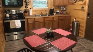 A compact kitchen with wooden cabinets, a microwave, a stove, and a sink. There is a dining table with red placemats and a potted plant.