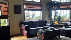 A living room with a brown leather sofa, a wooden coffee table, and a large window with a view of trees. - Comfortable Montana Country Home for Family Getaways (Arlee)