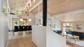 Un intérieur spacieux et moderne, doté d’un haut plafond, d’un coin repas avec des chaises transparentes, d’un espace salon avec un canapé et d’un escalier menant à un niveau supérieur.