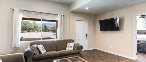 A spacious living room with a large window, a ceiling fan, a flat-screen TV mounted on the wall, and a wooden coffee table.