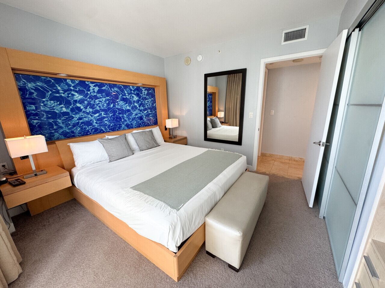 A hotel room with a large bed, a bedside table with a lamp, a bench, a large mirror, and a blue abstract painting on the wall.