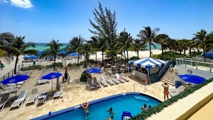 Zwembadgedeelte met ligbedden en parasols, een strand op de achtergrond en palmbomen.