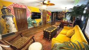Un salon chaleureux et accueillant, avec un canapé jaune, des fauteuils en bois, une batterie et un ventilateur de plafond.
